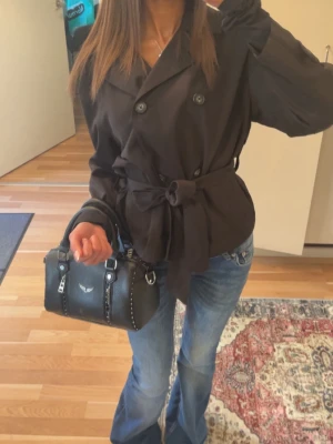 Svart trenchcoat med knytskärp - Stilren svart trenchcoat med dubbelknäppning och brett knytskärp i midjan. Jackan har klassisk krage och långa ärmar, perfekt för en trendig look. Passar snyggt till jeans eller klänning.