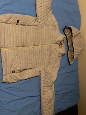 Ljusgrå pufferjacka med avtagbar huva - Snygg ljusgrå pufferjacka med quiltat mönster och avtagbar huva. Jackan har dragkedja framtill, två fickor med dragkedja och orange detaljer på dragkedjorna. Perfekt för kalla vinterdagar och riktigt skön att bära.