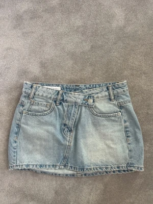 Blå jeanskjol från Bershka - Snygg ljusblå jeanskjol från Bershka med klassisk femficksdesign och bälteshällor. Kjolen har en kort passform och stängs med knapp och dragkedja framtill. Perfekt till vardags för en avslappnad look.