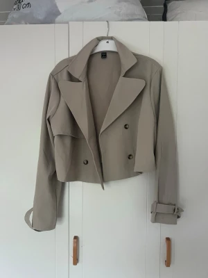 Beige cropped trenchcoat från shein - Stilren beige cropped trenchcoat från shein med dubbelknäppning och breda slag. Jackan har bälte vid ärmsluten och klassisk trenchcoat-design. Perfekt för dig som vill ha en modern twist på en klassiker. Köpte och har inte användning av den där av säljer💗