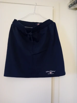 Mörkblå kortkjol från Grace Original - Mörkblå kortkjol från Grace Original med knyt i midjan och broderad logga nertill. Kjolen har en enkel och stilren design, perfekt för en avslappnad look. Tillverkad i mjukt material som känns bekvämt mot huden.