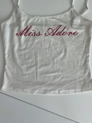 Vit linnetopp med text Miss Adore - Vit linnetopp med tunna axelband och tryckt texten 'Miss Adore' i vinrött framtill. Enkel och stilren design, med ställbara band, perfekt att matcha med jeans eller kjol. 