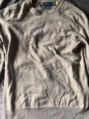Beige tröja från Polo Ralph Lauren - Klassisk beige tröja från Polo Ralph Lauren med rund halsringning och diskret broderad logga på bröstet. Tröjan är tunnstickad och har ribbade muddar vid ärmslut och nederkant. Perfekt för en stilren och enkel look.
