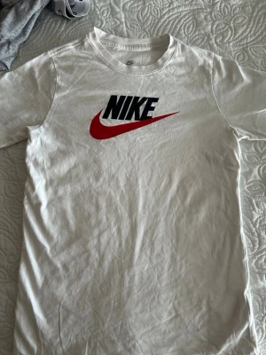 Vit Nike t-shirt med röd swoosh - Snygg vit t-shirt från Nike med klassisk svart logga och röd swoosh på bröstet. Tillverkad i mjuk bomull och har en rund halsringning. Perfekt för en sportig och avslappnad stil.