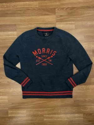 Mörkblå tröja från Morris Gent's - Mörkblå stickad tröja från Morris Gent's med röd text och detaljer. Tryck på bröstet med 'Morris Since 1968' och röda ränder vid ärmslut och nederkant. Rund halsringning och klassisk passform. Perfekt för dig som gillar stilrena och sportiga plagg.