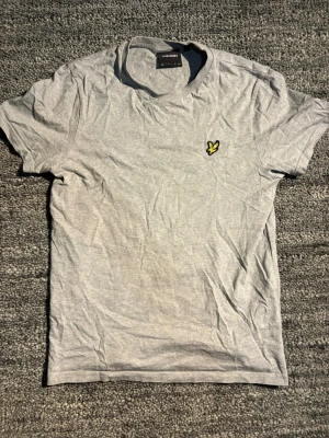 Grå t-shirt från Lyle & Scott - Snygg grå t-shirt från Lyle & Scott med klassisk gul logga på bröstet. T-shirten har rund hals och korta ärmar, tillverkad i mjuk bomull för en skön känsla. Perfekt basplagg till garderoben.