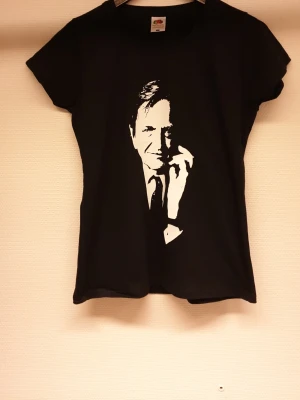 Olof Palme  t-shirt med tryck - Cool svart t-shirt från Fruit of the Loom med ett vitt tryck av Olof Palme på framsidan. Modellen är Lady Fit och har korta ärmar och rund hals. Perfekt för dig som vill sticka ut med en unik look.
