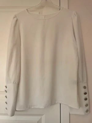 Vit blus med knappar på ärmarna - Stilren vit blus med rund halsringning och långa ärmar. Ärmsluten pryds av dekorativa knappar som ger en trendig touch. Perfekt att matcha med både jeans och kjol för en clean look. Materialet ser ut att vara mjukt och lätt, troligen polyester eller viskos.