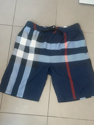 Burberry shorts - Snygga badbyxor från Burberry i marinblått med klassiskt rutmönster i vitt, svart, ljusblått och en röd rand. Elastisk midja och snabbtorkande material, perfekta för stranden eller poolen. Ikonisk design som sticker ut.