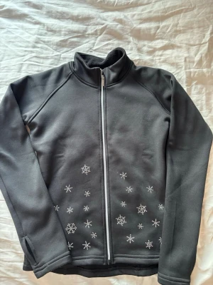 Svart konståknings jacka med snöflingor - Snygg svart konståknings jacka med hel dragkedja och hög krage. Jackan har vita snöflinge-mönster i nederkant både fram och bak, vilket ger en vintrig vibe. Tillverkad i ett stretchigt och mjukt material som passar perfekt för utomhusaktiviteter.varm och skön att ha på träning. Storlek 140 