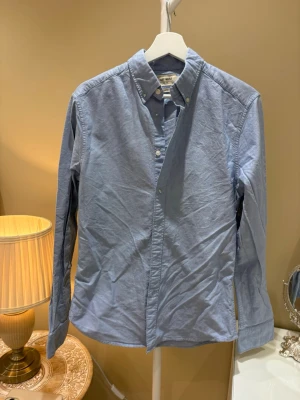 Blå skjorta från East West - Klassisk ljusblå skjorta från East West med button down-krage och slim fit passform. Skjortan har långa ärmar och knappar framtill. 