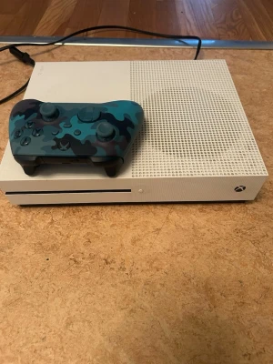 Xbox One S med blå kamouflagekontroll (Minecraft kommer med) - Säljer en vit Xbox One S med en unik blå kamouflage Xbox-kontroll. Konsolen har en stilren, rektangulär design med ventilerad ovansida och klassisk Xbox-logga. Kontrollern har ett snyggt blått camomönster och är tillverkad i plast. Spelet: Minecraft, kommer med.