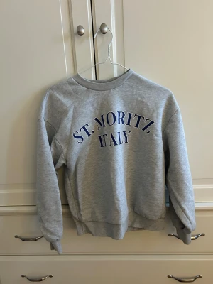 Grå sweatshirt St. Moritz Gina Tricot - Säljer en ljusgrå sweatshirt från Gina Tricot med blå text 'ST. MORITZ ITALY' på bröstet. Tröjan har rund halsringning, ribbade muddar vid ärmslut och nederkant samt en avslappnad passform. Perfekt för en chill och sportig look.