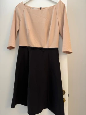 Beige och svart klänning Rinascimento - Snygg tvåfärgad klänning från Rinascimento med beige överdel och svart kjoldel. Klänningen har trekvartsärm, rund halsringning och dragkedja bak. Stilren design som passar perfekt till många tillfällen.
