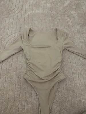 Beige långärmad body med fyrkantig ringning - Snygg beige body med lång ärm och fyrkantig ringning. Modellen har rynkade sidor som ger en smickrande passform. Tillverkad i ett stretchigt syntetmaterial som sitter tight mot kroppen. Perfekt att styla med jeans eller kjol.