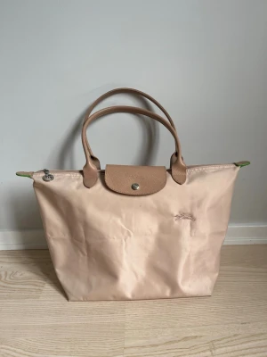 Baby Rosa Longchamp Le Pliage handväska - Säljer en klassisk baby rosa Longchamp Le Pliage handväska i nylon med detaljer i ljus rosa läder. Väskan har dubbla handtag, tryckknapp och dragkedja. Den har använts mycket! 😊