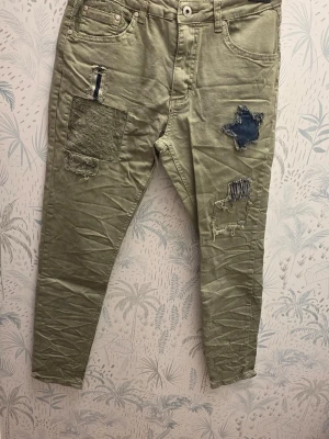 Cat & Co Jeans, size 36 - Säljer ett par olivgröna jeans med flera coola patchar och slitningar framtill. Jeansen har smal passform, klassiska fickor och unika detaljer som spets och denimlappar. Perfekta för dig som gillar en avslappnad och trendig stil.