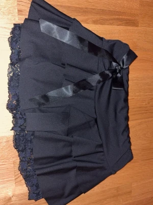 Svart volangkjol med spets och rosett - Supersöt svart kortkjol med volanger, spetskant och en dekorativ rosett framtill. 