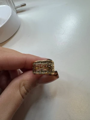Bred guldfärgad ring med mönster - Snygg bred ring i guldfärg med ett klassiskt grekiskt mönster och glittrande stenar längs kanterna. Ringen har en chunky känsla och passar perfekt för dig som gillar statement-smycken. Materialet ser ut att vara metall med guldfinish.