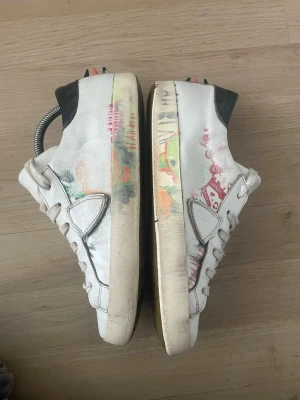 Vita phillipe models  - Unika vita sneakers med färgstänk i grönt, orange och rött. Skorna har svarta detaljer vid hälen och vita snören. Låg modell med klassisk siluett och coola grafiska inslag på sidorna. Tillverkade i läder med gummisula. Perfekta för dig som vill sticka ut.