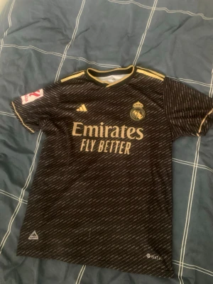 Real Madrid svart matchtröja Adidas M - Säljer en svart Real Madrid matchtröja från Adidas med gulddetaljer och klubbmärke på bröstet. Tröjan har 'Emirates Fly Better' tryckt framtill och är tillverkad i lätt och ventilerande AEROREADY-material. Perfekt för fotboll eller träning.