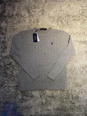 Grå Ralph Lauren tröja - Säljer denna klassiska kabelstickade Ralph Lauren tröjan i grått med svart logga. Storleken är M fast passar närmre S-XS enligt mig, hör gärna av dig vid frågor om t.ex. mått, bilder etc!
