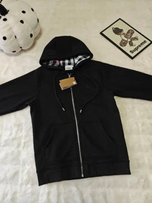 Svart hoodie med dragkedja Burberry - Svart hoodie med dragkedja från Burberry. Klassisk huva med dragsko och insida i Burberrys rutiga mönster. Två fickor framtill och ribbade muddar. Perfekt för en stilren och trendig look.