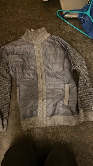 Moncler liknade Grå cardigan - Snygg moncler liknande  grå jacka från okänt märke med stickade ärmar och krage, quiltad polyester på framsidan och baksidan samt dragkedja framtill. Insidan har ett mönstrat fleecefoder i svart och vitt. Perfekt för dig som gillar en chill och sportig look. Den är super mysig inuti 