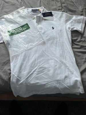 Vit pikétröja från Polo Ralph Lauren - (SPRILLANS NY)Klassisk vit pikétröja från Polo Ralph Lauren med krage, knappar och den ikoniska broderade loggan på bröstet. Tröjan har korta ärmar och är tillverkad i mjuk bomull som känns skön mot huden. Perfekt till jeans eller shorts för en clean look.