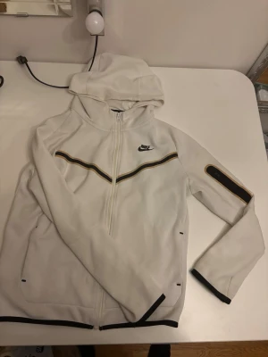 Vit Nike tech kofta med dragkedja - Säljer en vit hoodie från Nike med hel dragkedja framtill och svart Nike-logga på bröstet. Hoodien har svarta och gula detaljer längs bröstet och ärmslut samt en ficka med dragkedja på ena ärmen. Perfekt för en sportig och clean look.