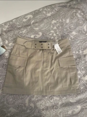 Beige kortkjol med bälte och fickor - Snygg beige kortkjol med bredt bälte och stora cargofickor på sidorna. Kjolen har en rak passform och är tillverkad i ett slitstarkt bomullsmaterial. Perfekt för en trendig och avslappnad look.