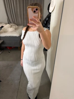 Vit långklänning med offshoulder - Säljer en elegant vit långklänning med offshoulder-design och smala axelband. Klänningen har ett plisserat tyg som ger en snygg struktur och följer kroppen på ett smickrande sätt. Perfekt för dig som vill ha en stilren och modern look.