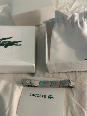 Silverfärgat armband från Lacoste - Stilrent armband från Lacoste i silverfärgad metall med klassisk krokodil-logga på spännet. Länkarna har en snygg meshstruktur som ger en modern känsla. Kommer i original ask och med tygpåse, perfekt för dig som gillar minimalistisk och sportig design.