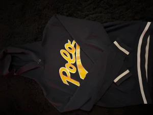 Polo Ralph Lauren Hoodie - Mörkblå hoodie med stor gul Polo-logga på bröstet och vita ränder på ärmslut och nederkant. Klassisk huva och mjukt material som känns skönt mot huden. Perfekt för dig som gillar streetstyle och vill ha något som sticker ut.