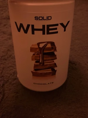 Solid Nutrition Whey Chocolate - Säljer en burk Solid Nutrition Whey proteinpulver med chokladsmak. Förpackningen är vit med svart text och har en bild på chokladbitar. Innehåller 750g och är laktosfri. Perfekt för dig som tränar och vill ha extra protein. Det är mer än halva kvar