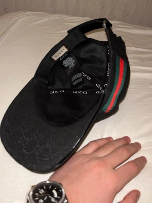 Svart Gucci keps med GG-mönster - Snygg svart keps från Gucci med klassiskt GG-monogram i tyget och justerbart spänne bak i läder. På sidan finns den ikoniska gröna och röda randen. Kepsen är tillverkad i ett slitstarkt tyg och har detaljer i läder för en lyxig känsla.