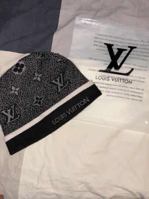 Svart och grå Louis Vuitton mössa - Exklusiv svart och grå mössa från Louis Vuitton med klassiskt monogrammönster och broderad logga framtill. Mössan har en bred, ljusgrå kant och är tillverkad i ett mjukt, stickat material som känns lyxigt och bekvämt. Perfekt för dig som vill ha en stilren accessoar med premiumkänsla.