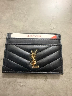 Svart YSL kortfodral i läder - Snyggt svart kortfodral från YSL i quiltat läder med guldigt logotypemblem framtill. Perfekt för att hålla koll på dina kort med stil. Smidig och tunn design som enkelt får plats i fickan.