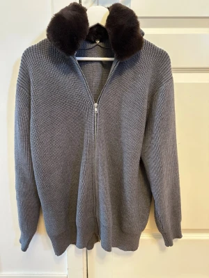 Grå stickad kofta med pälskrage - Säljer en grå stickad kofta med dragkedja och avtagbar brun fusk-pälskrage. Koftan har ribbstickade muddar och en avslappnad passform. Perfekt för dig som vill ha något både mysigt och stilrent.