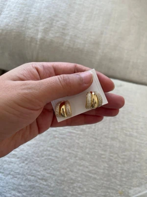 Chunky guldfärgade örhängen - Snygga chunky örhängen i guldpläterad med en bred, rundad form. Örhängena har en mittdel i blank metall och sidorna är täckta av små gnistrande stenar som ger extra bling. Perfekta för dig som gillar statement-smycken och vill sticka ut. Rostfritt stål. Vattentålig 