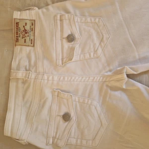 Vita bootcut jeans från True Religion - Säljer ett par vita True Religion jeans som är bootcut som är lågmidjade. Innerbenslängden är 80cm och midjemåttet rakt över 36cm. Nyköpta kostar 1300 kr