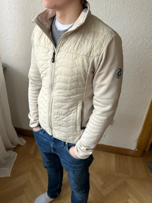 Beige quiltad jacka från Pelle p - Snygg beige jacka från Pelle p  med quiltad front och rygg samt mjuka ärmar. Jackan har hög krage, dragkedja framtill och en diskret logga på ärmen. Perfekt för lager-på-lager och passar till jeans eller träningskläder.