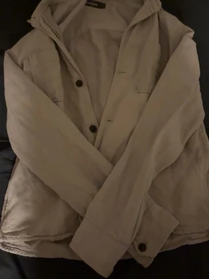 Beige overshirt från Dressmann - Snygg beige overshirt från Dressmann i storlek S. Jackan har en klassisk krage, bröstficka och knäppning med svarta knappar. Tillverkad i mjukt bomullsmaterial, perfekt att slänga på över en t-shirt eller tröja. Enkel och stilren design som funkar till jeans.