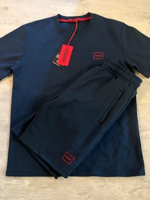 Mörkblå bodyset från Hugo - Snyggt mörkblått bodyset från Hugo med matchande t-shirt och shorts. Båda delar har diskret röd Hugo-logga på bröstet och benet. Materialet är mjukt och skönt bomull, perfekt för en avslappnad och stilren look. ! DET FINNS STORLEKAR S-M-L-XL !