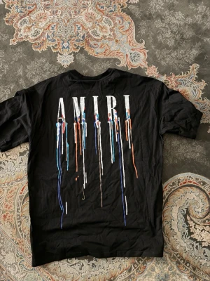 Svart AMIRI t-shirt med färgstänk - Säljer en svart t-shirt från AMIRI med stort tryck på ryggen där AMIRI står i vitt och färgstänk i blått, orange och vitt rinner ner. Klassisk passform och rund hals. Perfekt för dig som gillar streetwear och vill sticka ut.