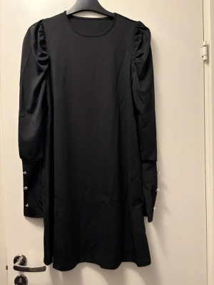 Svart klänning med puffärmar - Stilren svart klänning med rund halsringning och långa puffärmar. Ärmarna har dekorativa knappar längs underarmen som ger en extra detalj. Klänningen har en rak passform och är i kort modell.