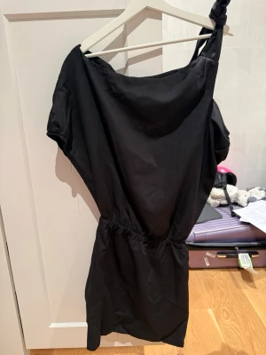 Svart oneshoulder kortklänning - Svart kortklänning med oneshoulder-design och knyt på axeln. Klänningen har en avslappnad passform med resår i midjan som ger en snygg siluett. Perfekt för dig som gillar en stilren och trendig look. Originalpris ca 340kr