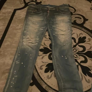 Dsquared2 ljusblå distressed jeans - Säljer ett par ljusblå jeans från Dsquared2 med slitna detaljer och färgstänk för en riktigt edgy look. Jeansen har klassisk femficksmodell och låg midja. Perfekta för dig som gillar streetwear och vill sticka ut lite extra.