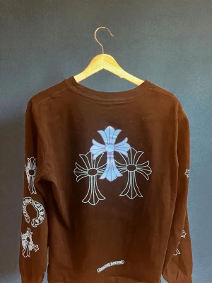  tröja Chrome Hearts - Cool brun långärmad tröja från Chrome Hearts med stora vita och blå kors på ryggen och ärmarna. Bröstficka med tryck och detaljerade loggor på ärmar och fram. Skön passform och crewneck, perfekt för dig som gillar streetwear och unika prints.