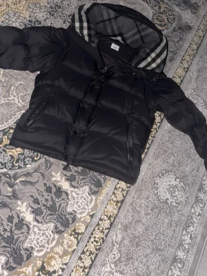 Den kända burberry jackan - Säljer en svart pufferjacka med dragkedja och knappar framtill. Jackan har två sidofickor med dragkedja och en stor huva med grått och vitt rutigt foder. Perfekt för kalla dagar och riktigt skön att bära. Kan även dra av armarna så de bildas till en vest.